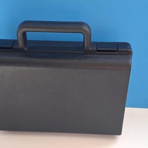 May include: A black briefcase with a handle on top. The rectangular case has a smooth, matte finish and a secure closure. The handle is also black and appears sturdy. The background is a solid blue.
