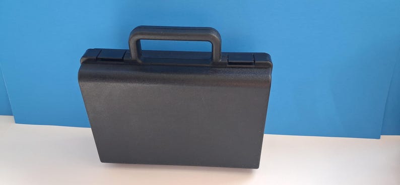 May include: A black briefcase with a handle on top. The rectangular case has a smooth, matte finish and a secure closure. The handle is also black and appears sturdy. The background is a solid blue.