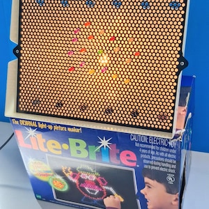 Könnte beinhalten: Ein Vintage Lite-Brite-Spielzeug mit einem schwarzen Bildschirm mit zahlreichen kleinen Löchern, der von einem warmen Licht beleuchtet wird. Auf der Box steht "The ORIGINAL light-up picture maker!" und zeigt ein farbenfrohes Bild. Ein klassisches Kreativspielzeug.