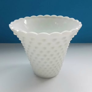 Puede incluir: Un jarrón blanco con diseño de hobnail y borde festoneado. El jarrón tiene forma acampanada y una superficie texturizada con pequeñas protuberancias espaciadas uniformemente. El fondo es un degradado de blanco y azul.