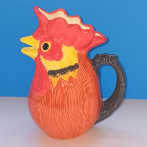May include: A ceramic pitcher shaped like a rooster. The pitcher is primarily orange with a red comb and wattle, yellow beak and neck feathers, and a black handle. The pitcher has a glossy finish and is set against a blue background.