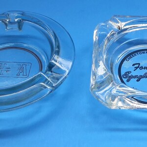 May include: Two clear glass ashtrays. The round one features the letters RF A in a rectangular box. The square one displays the text Fontana's Spaghetti House, Cheektowaga, 822-7915, and 822-4444 printed in black ink on the bottom surface.