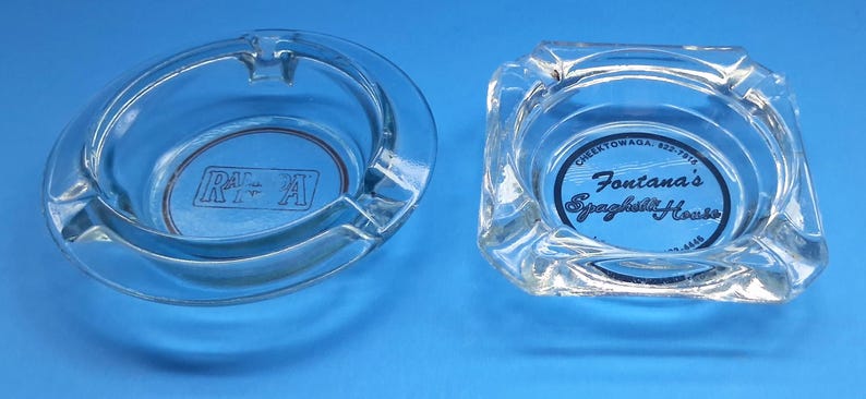 May include: Two clear glass ashtrays. The round one features the letters RF A in a rectangular box. The square one displays the text Fontana's Spaghetti House, Cheektowaga, 822-7915, and 822-4444 printed in black ink on the bottom surface.