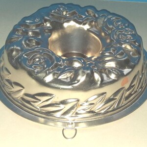 Puede incluir: Un molde para pasteles circular de metal color plateado con un patr&oacute;n en relieve de flores y hojas en la superficie superior. El molde tiene un tubo central hueco y una peque&ntilde;a anilla de metal en el borde exterior.