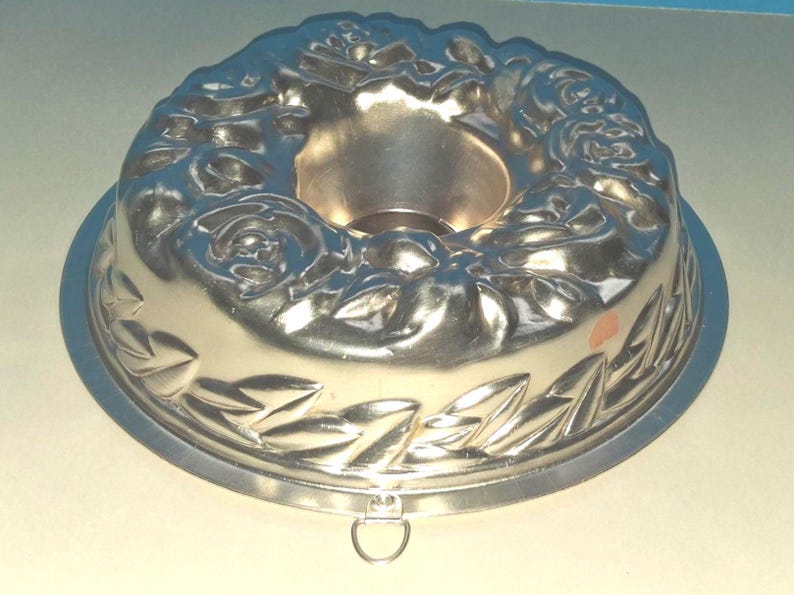 Puede incluir: Un molde para pasteles circular de metal color plateado con un patr&oacute;n en relieve de flores y hojas en la superficie superior. El molde tiene un tubo central hueco y una peque&ntilde;a anilla de metal en el borde exterior.