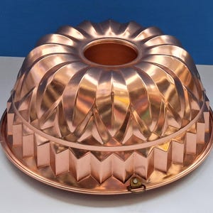 May include: A copper-colored bundt cake pan with a metallic finish. The pan features a central tube, a fluted exterior with vertical ridges, and a distinct zigzag pattern along the base rim. A small metal hanging loop is attached to the bottom edge.