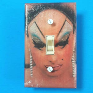 May include: A light switch cover with a colorful portrait of a person with heavy makeup and a light switch in the center. The text "love" is visible at the bottom of the image.