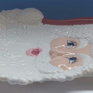 May include: A white ceramic Santa Claus face-shaped dish with a red hat. The dish has a detailed design of Santa's beard and face, with blue eyes and a pink nose. The dish is likely for decorative or serving purposes.
