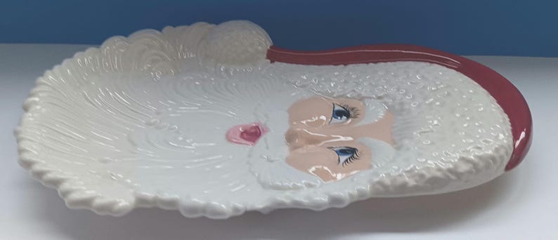May include: A white ceramic Santa Claus face-shaped dish with a red hat. The dish has a detailed design of Santa's beard and face, with blue eyes and a pink nose. The dish is likely for decorative or serving purposes.