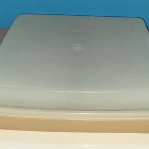 Könnte beinhalten: Ein quadratischer, beigefarbener Kunststoff-Aufbewahrungsbehälter mit einem durchscheinenden, halb-opaken Deckel. Die Basis verfügt über integrierte Seitengriffe und einen flachen Boden, während der Deckel sicher oben aufliegt und eine Einheit bildet.