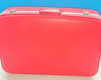 Può includere: Una valigia rettangolare rosso brillante con un esterno liscio e a tinta unita. Il bagaglio presenta una maniglia rossa montata sulla parte superiore, un bordo bianco lungo la chiusura e due chiusure metalliche color argento.