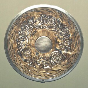 Puede incluir: Un molde de horneado redondo de metal que presenta un patr&oacute;n en relieve detallado de rosas y hojas a lo largo de la base interior. El centro del molde es liso y circular, rodeado por el dise&ntilde;o floral, con un acabado plateado pulido.