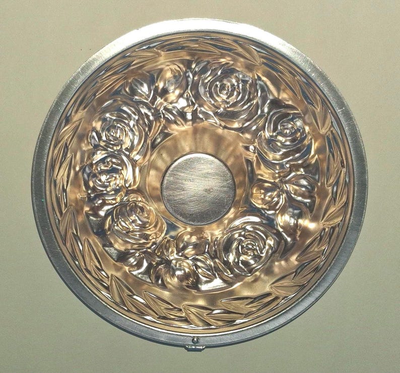 Puede incluir: Un molde de horneado redondo de metal que presenta un patr&oacute;n en relieve detallado de rosas y hojas a lo largo de la base interior. El centro del molde es liso y circular, rodeado por el dise&ntilde;o floral, con un acabado plateado pulido.