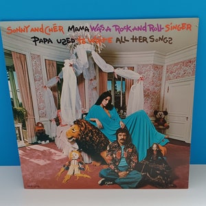 Könnte beinhalten: Albumcover von Sonny und Cher's "Mama Was a Rock and Roll Singer, Papa Used to Write All Her Songs?" mit einem Foto des Duos. Cher trägt ein türkisfarbenes Kleid und Sonny eine Blumenjacke. Der Hintergrund ist ein Raum mit rosa Tapeten.