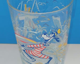 May include: Clear glass tumbler featuring a cartoon character skiing. The tumbler has a blue rim and a colorful design of a cartoon character in red and white striped pants, skiing on a white background with a blue sky.
