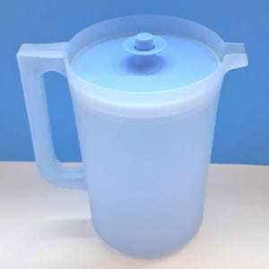 May include: A light blue plastic pitcher with a matching lid and a side handle. The pitcher features a cylindrical body, a small pouring spout on the rim, and a circular knob on the center of the lid for easy removal. The design is simple and functional.