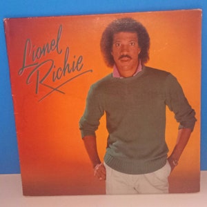 May include: Album cover featuring Lionel Richie against an orange and red gradient background. The album title "Lionel Richie" is written in a cursive font. Richie is wearing a green sweater and light-colored pants.