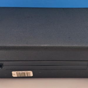 May include: A closed, black rectangular case with a textured surface. The case has two hinges and a latch on the front. A small white sticker with a barcode is affixed to the front of the case. The case is sitting on a white surface.
