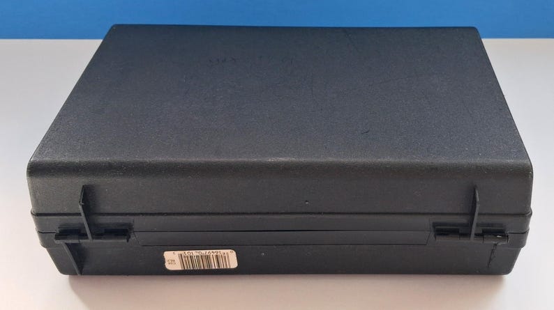 May include: A closed, black rectangular case with a textured surface. The case has two hinges and a latch on the front. A small white sticker with a barcode is affixed to the front of the case. The case is sitting on a white surface.