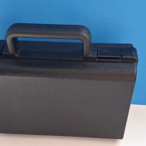 May include: A black, rectangular briefcase with a handle and two latches. The case is made of a textured material and sits on a white surface against a blue backdrop. The handle is arched and made of the same black material.