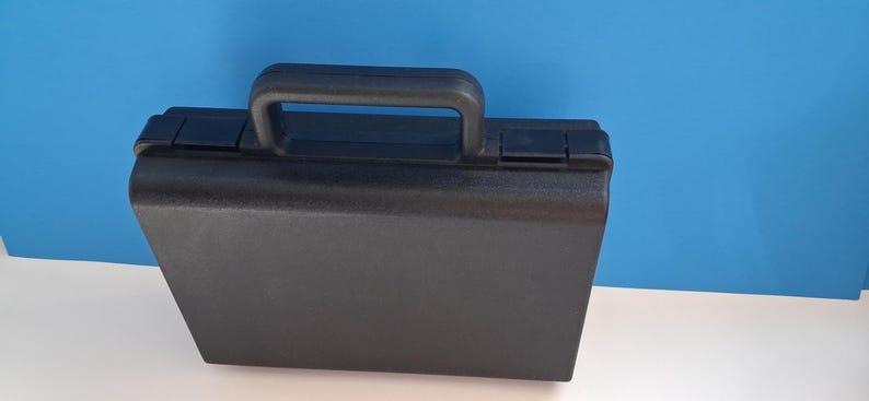 May include: A black, rectangular briefcase with a handle and two latches. The case is made of a textured material and sits on a white surface against a blue backdrop. The handle is arched and made of the same black material.