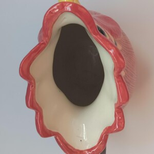 May include: A ceramic pitcher in the shape of a rooster, viewed from above. The pitcher features a red comb and wattle, a yellow beak, and a black handle. The interior is dark brown, and the rim is outlined in red.