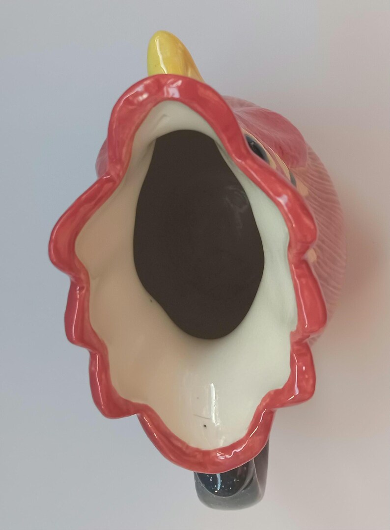 May include: A ceramic pitcher in the shape of a rooster, viewed from above. The pitcher features a red comb and wattle, a yellow beak, and a black handle. The interior is dark brown, and the rim is outlined in red.