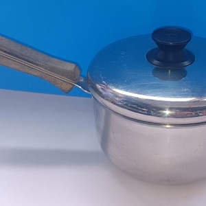 May include: A small, stainless steel saucepan with a lid and a handle. The lid has a black knob. The handle is silver with a brown grip. The saucepan is on a white surface with a blue background.