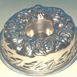 Puede incluir: Un molde circular de metal color plata para pasteles tipo bundt, con un dise&ntilde;o en relieve de rosas florecientes alrededor del borde superior y un patr&oacute;n decorativo de hojas grabado en el borde inferior, con un tubo central hueco.
