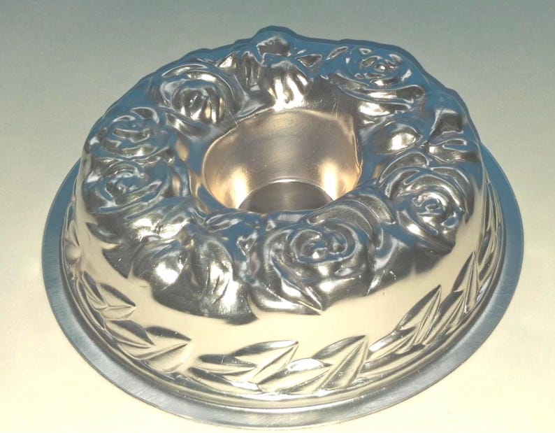 Puede incluir: Un molde circular de metal color plata para pasteles tipo bundt, con un dise&ntilde;o en relieve de rosas florecientes alrededor del borde superior y un patr&oacute;n decorativo de hojas grabado en el borde inferior, con un tubo central hueco.