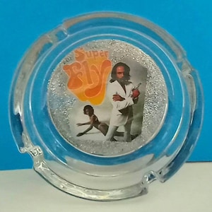 May include: A clear glass ashtray with a silver glitter background and a picture of a man in a white suit with the words "Super Fly" in orange and yellow letters.