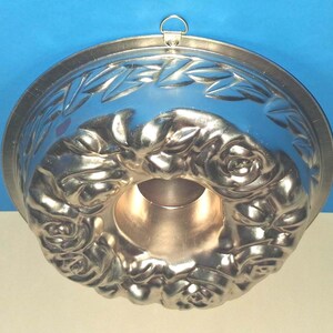 Puede incluir: Un molde para pasteles redondo de metal con un patr&oacute;n detallado en relieve de rosas y hojas en la superficie interior. El molde tiene un tubo central hueco y un peque&ntilde;o lazo de metal unido al borde superior para colgarlo.