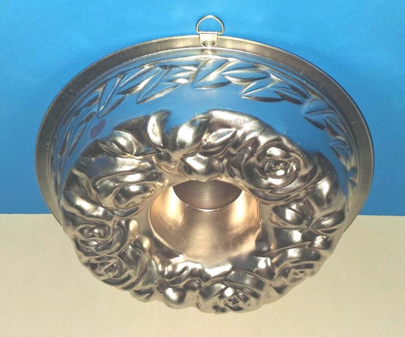 Puede incluir: Un molde para pasteles redondo de metal con un patr&oacute;n detallado en relieve de rosas y hojas en la superficie interior. El molde tiene un tubo central hueco y un peque&ntilde;o lazo de metal unido al borde superior para colgarlo.