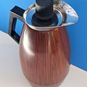 May include: A thermal carafe featuring a brown wood-grain patterned body, a black plastic handle, and a silver-toned metal rim. The top includes a black circular lid with a ridged knob for gripping, and a small pouring spout on the side.