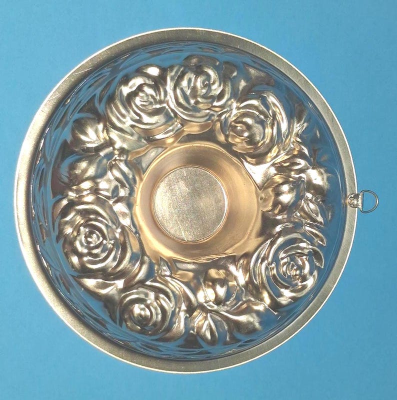 Puede incluir: Un molde de hornear redondo, met&aacute;lico y de color cobre, con un patr&oacute;n floral en relieve detallado de rosas alrededor de una hendidura circular central. El molde tiene una superficie lisa y reflectante e incluye una peque&ntilde;a anilla.