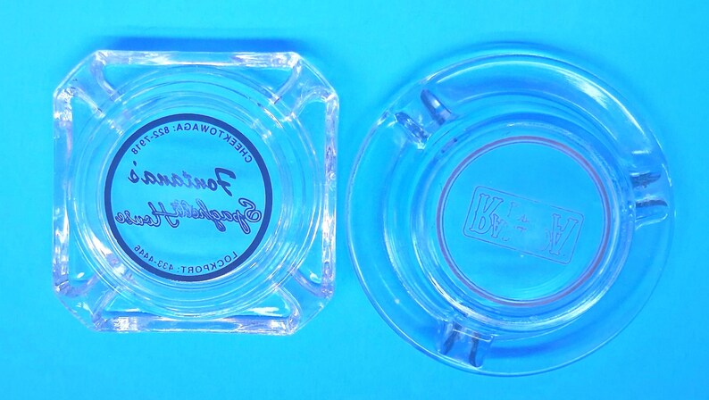 May include: A square clear glass ashtray with the text Fontana's Spaghetti House, Cheektowaga: 822-7918, Lockport: 433-4445 printed in black, and a round clear glass ashtray with the letters K and V separated by a star symbol etched in the center.