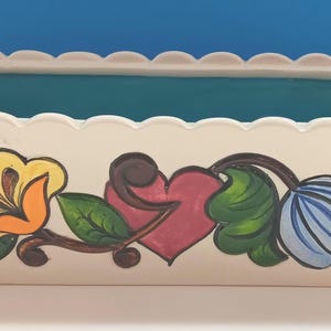 Könnte beinhalten: Cremefarbener rechteckiger Pflanztopf mit gewelltem Rand und türkisfarbenem Innenraum. Der Pflanztopf ist mit handgemalten Blumen- und Herzmotiven in Gelb, Orange, Blau und Grün verziert. Ein dekoratives Stück.