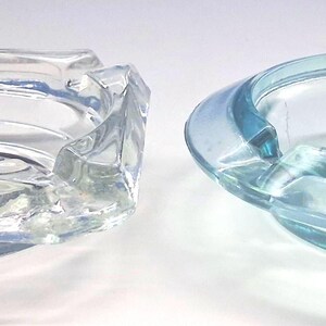 May include: Two glass ashtrays are displayed side by side. The left ashtray is clear, square-shaped with rounded corners and features the text TOWADA 507. The right ashtray is circular, light blue in color, and features two notches for resting items.