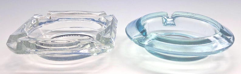 May include: Two glass ashtrays are displayed side by side. The left ashtray is clear, square-shaped with rounded corners and features the text TOWADA 507. The right ashtray is circular, light blue in color, and features two notches for resting items.