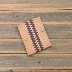 May include: A rectangular wooden cutting board with a light brown surface and a dark brown, zig-zag pattern down the center. The board is made of multiple wood pieces and sits on a wooden surface.