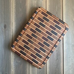 May include: Rectangular wooden cutting board with a brick-like pattern. The board features alternating rows of light and dark wood, with accents of dark brown and black. A groove runs around the edge of the board.