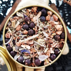 May include: A close-up of a round tin filled with a blend of dried herbs, including juniper berries, sage, and other botanicals. The tin is gold-colored and has a label that reads "The Magick Cabinet".