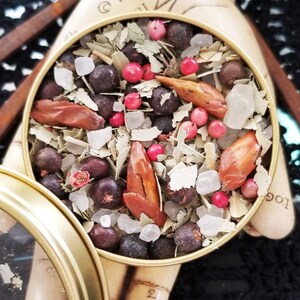 May include: A gold metal tin filled with a blend of herbs, spices, and crystals. The tin has a clear lid and the text "The Magick Cabinet&trade;" is printed on the bottom.