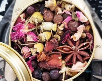 May include: A round metal tin filled with a blend of dried herbs, flowers, and spices. The tin is open and the contents are visible. The herbs and spices include rose petals, chamomile flowers, juniper berries, star anise, and other dried botanicals. The tin is labeled "The Magick Cabinet™".