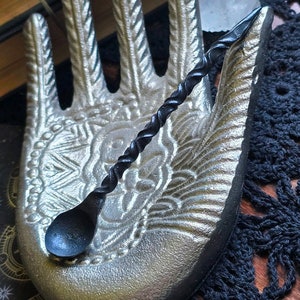 May include: A black, twisted, wooden spoon with a rounded bowl sits in a silver, hand-shaped dish. The dish has a detailed design of a hand with fingers and a palm.