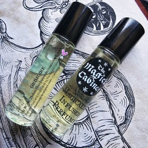 May include: Two small glass bottles with black roll-on tops filled with a clear liquid. The bottles are labeled "The Magick Cabinet" and "Grimoire" with a pink heart and stars. The bottles are on a white surface with a black and white illustration of a mythical creature.
