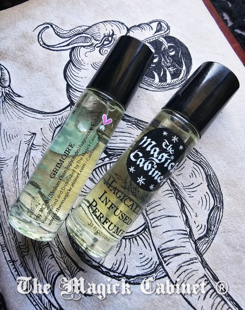 May include: Two small glass bottles with black roll-on tops filled with a clear liquid. The bottles are labeled "The Magick Cabinet" and "Grimoire" with a pink heart and stars. The bottles are on a white surface with a black and white illustration of a mythical creature.