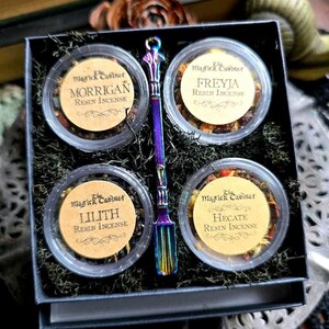 May include: A black box set containing four small, clear containers of resin incense labeled "Morrigan", "Freyja", "Lilith", and "Hecate". A multi-colored spoon rests between the containers. The box is lined with green moss and includes the text "The Magick Cabinet".