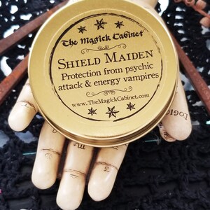 May include: Gold metal tin with a black label that reads "The Magick Cabinet" and "SHIELD MAIDEN Protection from psychic attack & energy vampires www.TheMagickCabinet.com". The label is decorated with five-pointed stars.