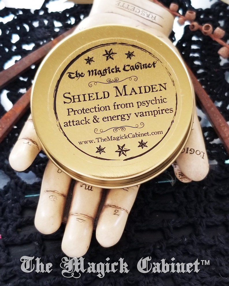 May include: Gold metal tin with a black label that reads "The Magick Cabinet" and "SHIELD MAIDEN Protection from psychic attack & energy vampires www.TheMagickCabinet.com". The label is decorated with five-pointed stars.
